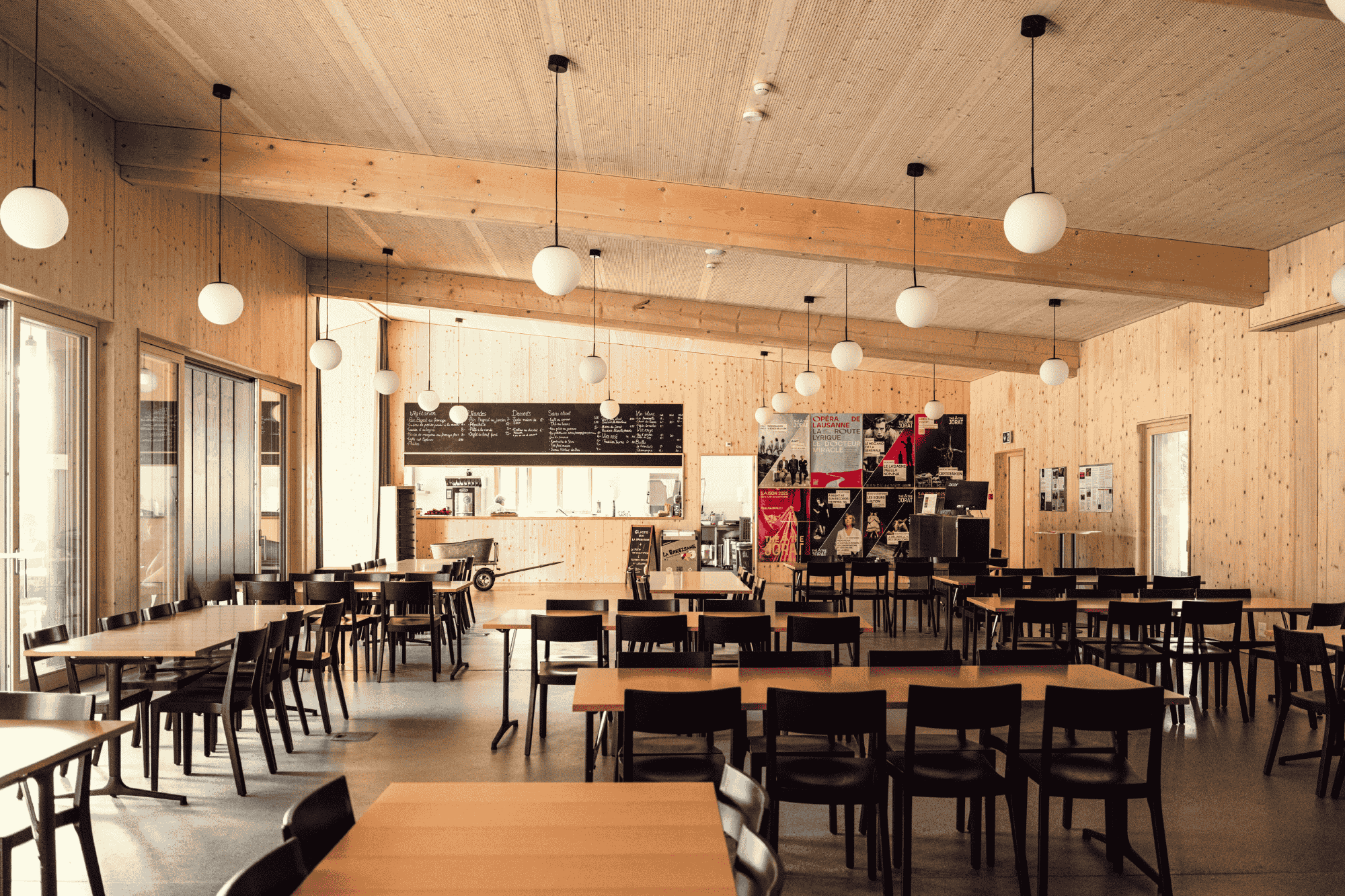 Intérieur du Pavillon du Théâtre du Jorat montrant des tables, des chaises, la cuisine au fond, des affiches et des luminaires.